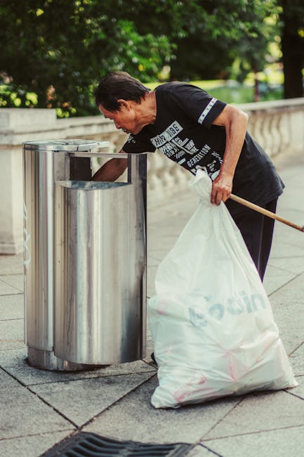 A middle-aged man with dark hair, dressed in a black T-shirt with white text, is seen disposing of rubbish into a cylindrical stainless steel public waste bin on a paved sidewalk. He is bending forwar