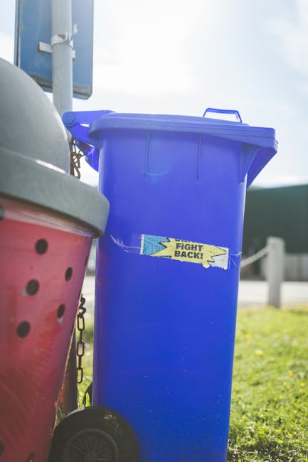 A middle-aged man with dark hair, dressed in a black T-shirt with white text, is seen disposing of rubbish into a cylindrical stainless steel public waste bin on a paved sidewalk. He is bending forwar